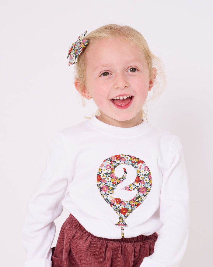 A 2 year old girl wearing a 2nd birthday white t-shirt with a balloon and number 2 on it made from Liberty Floral print fabric.