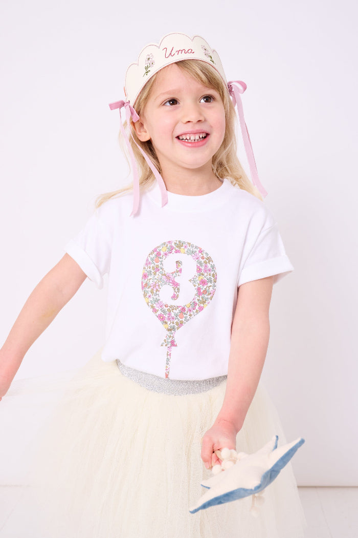 A 3 year old girl wearing a 3rd birthday white t-shirt with a balloon and number 3 on it made from Liberty Floral print fabric.