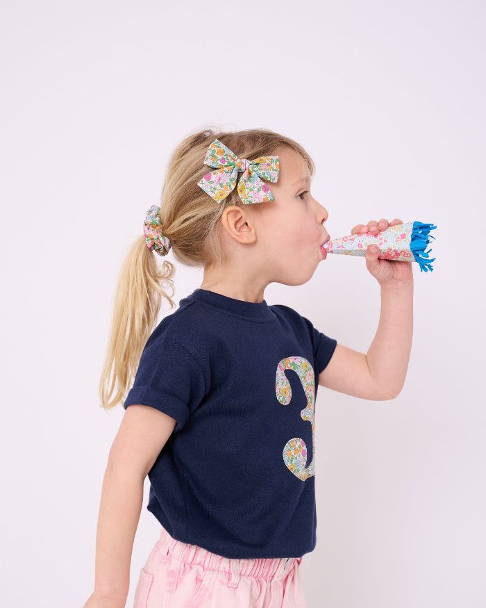 A 3 year old birthday girl wearing a Magnificent Stanley kids Number Navy T-Shirt in Poppy Forest Liberty Print. She wears a matching liberty scrunchie and bow clip.