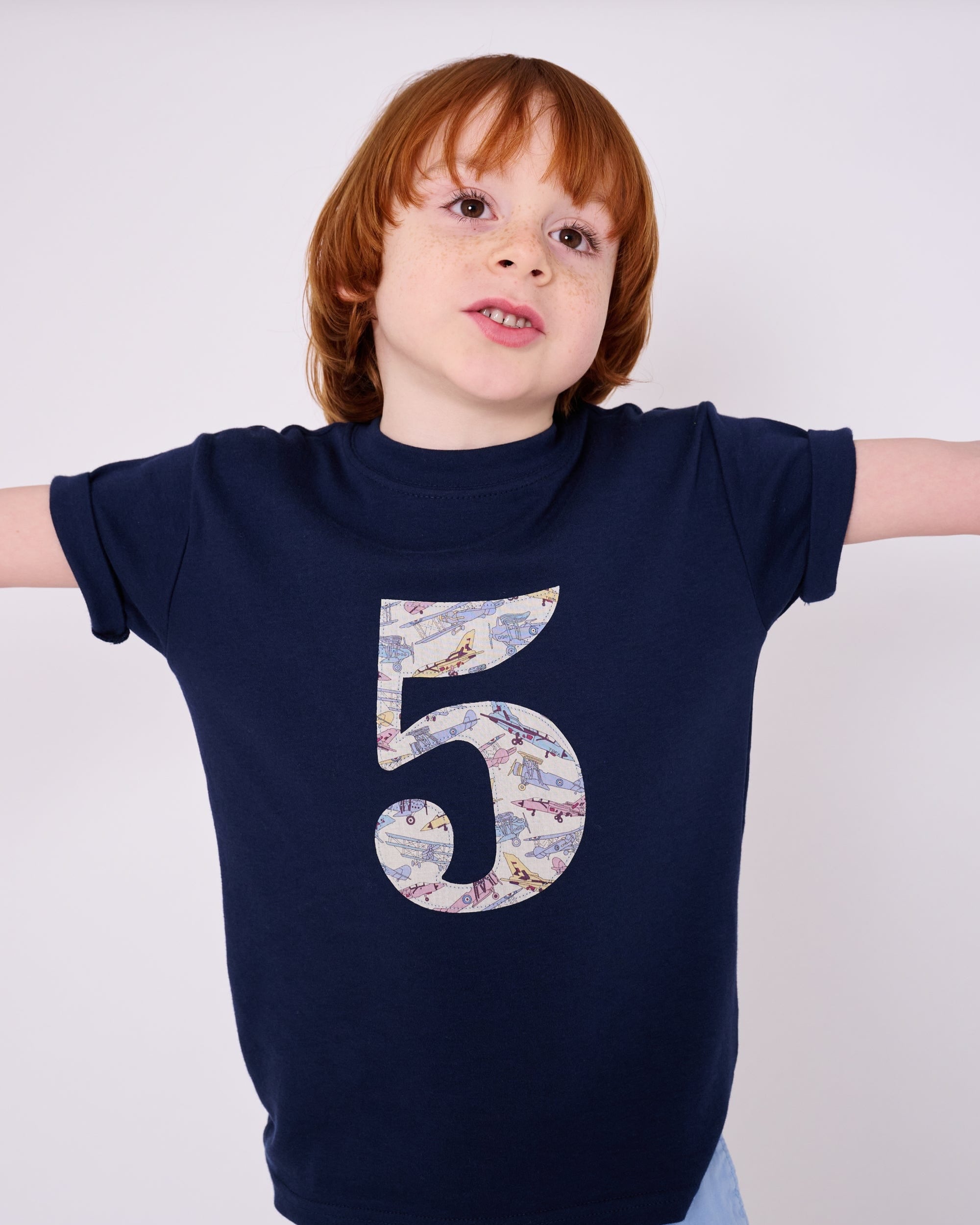 A 5 year old boy wearing a navy cotton t-shirt with a number 5 sewn on the front made from Tom's Jet plane Liberty fabric.