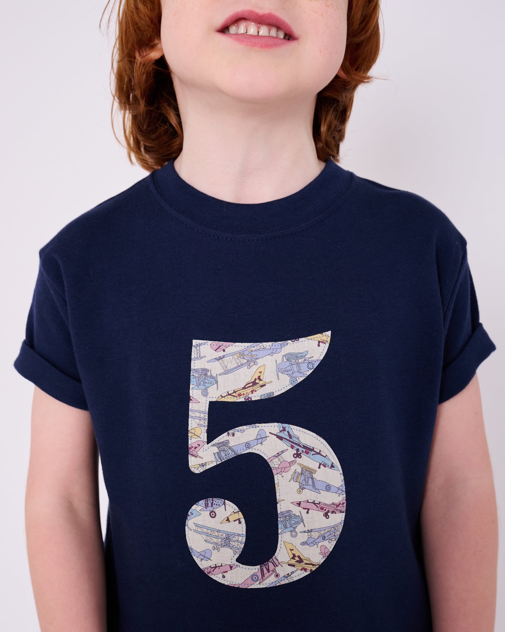 A 5 year old boy wearing a navy cotton t-shirt with a number 5 sewn on the front made from Tom's Jet plane Liberty fabric.