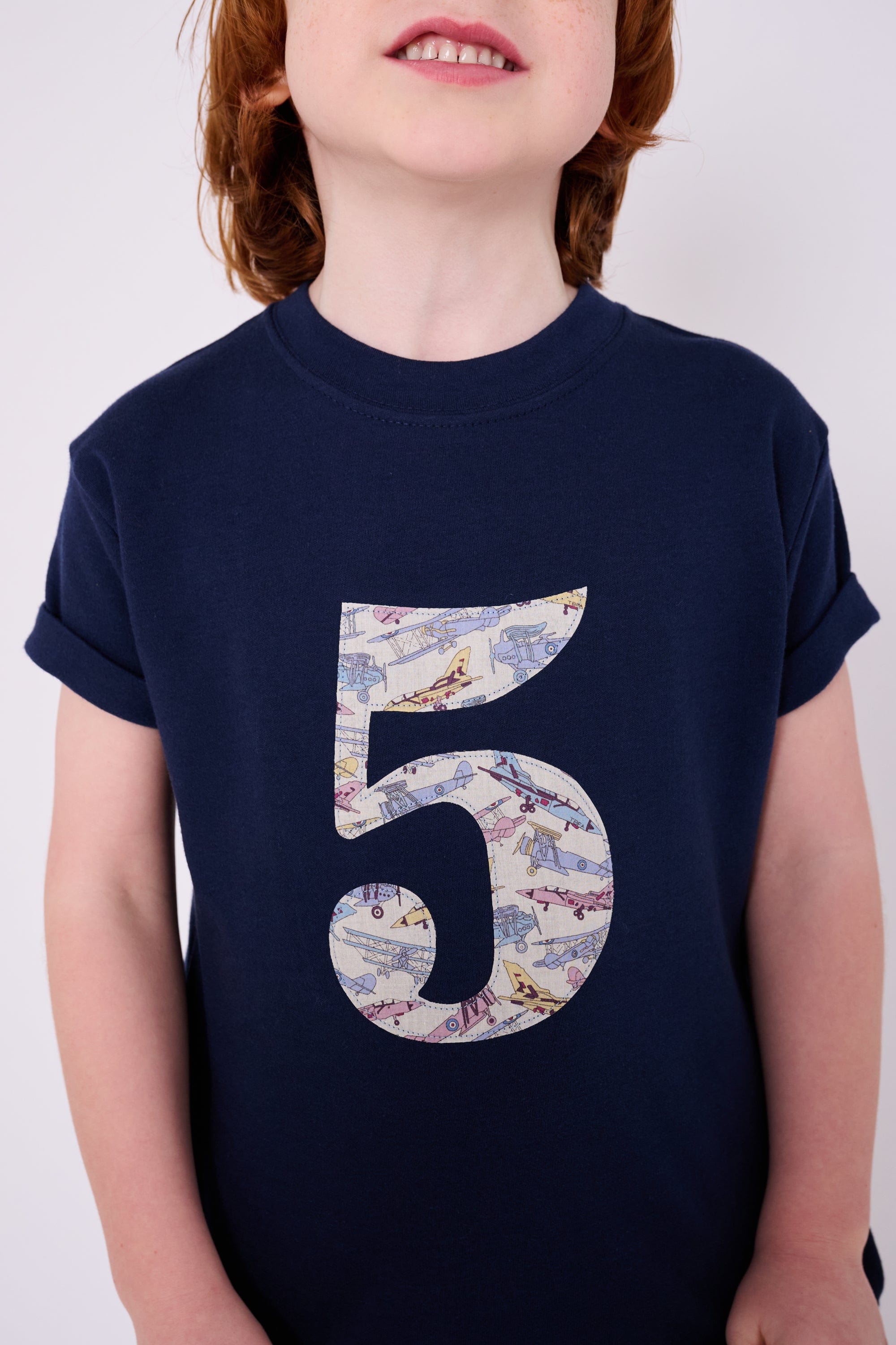 A 5 year old boy wearing a navy cotton t-shirt with a number 5 sewn on the front made from Tom's Jet plane Liberty fabric.
