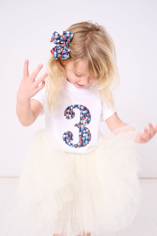A 3 year old birthday girl wearing a tutu skirt and a number 3 white cotton birthday T-Shirt in Ethan's Party balloon Liberty Print.