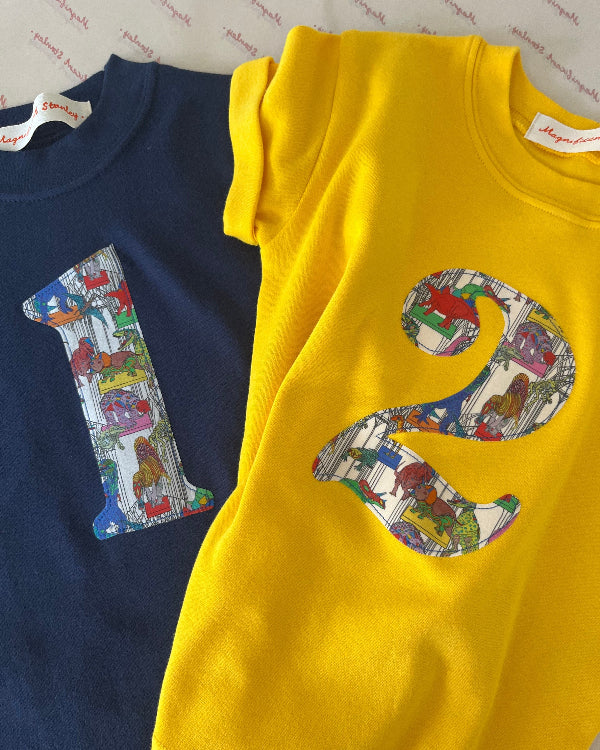 Navy and yellow kids t-shirts with a colourful number 1 and 2 design in Liberty dinosaur print and light blue shorts on a white background
