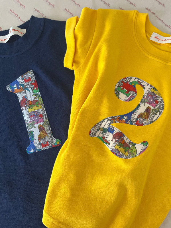 Navy and yellow kids t-shirts with a colourful number 1 and 2 design in Liberty dinosaur print and light blue shorts on a white background