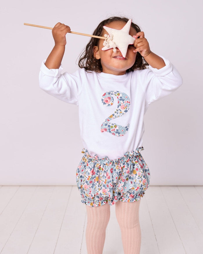 A child wearing a white t-shirt with the number 2 in Betsy Grey Liberty Print, paired with floral bloomers, holding a wand and standing against a white background.