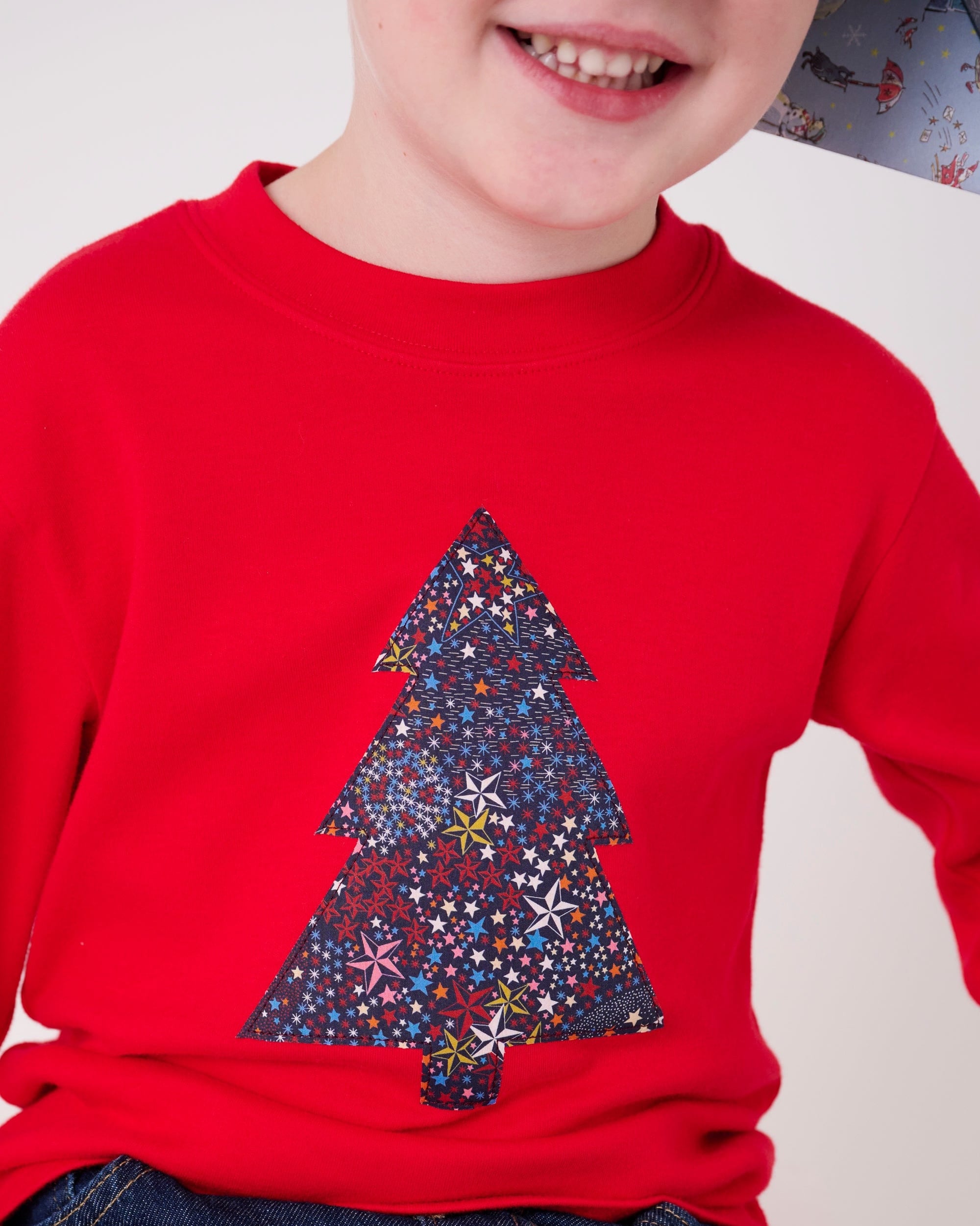 A 4 year old boy wearing a festive red cotton t-shirt with a christmas tree made from Liberty star Adelajda's Wish fabric appliquéd on the front.