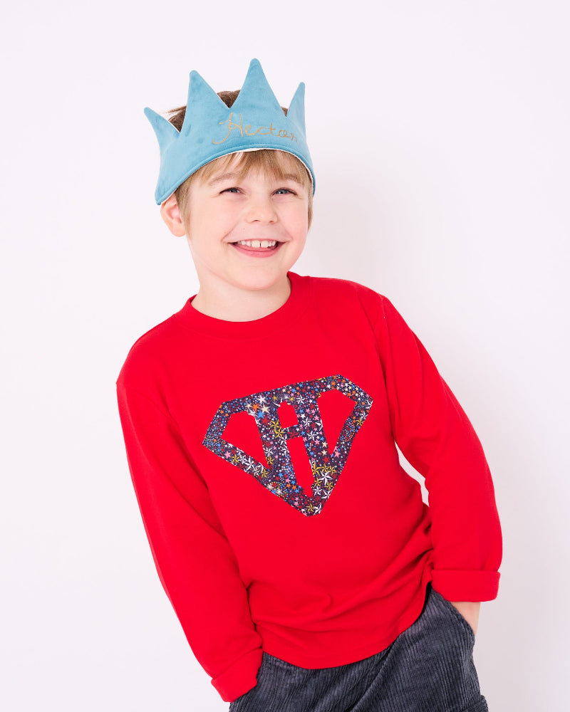 A smiling 5 year old boy wearing a festive red cotton personalised t-shirt with his initial in a superhero emblem Liberty star Adelajda's Wish fabric appliquéd on the front.