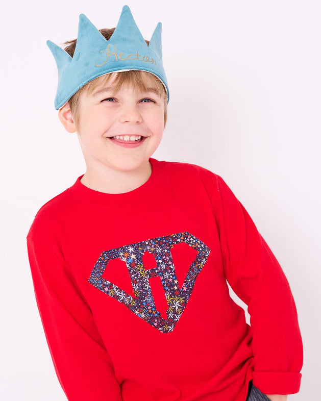 A smiling 5 year old boy wearing a festive red cotton personalised t-shirt with his initial in a superhero emblem Liberty star Adelajda's Wish fabric appliquéd on the front.