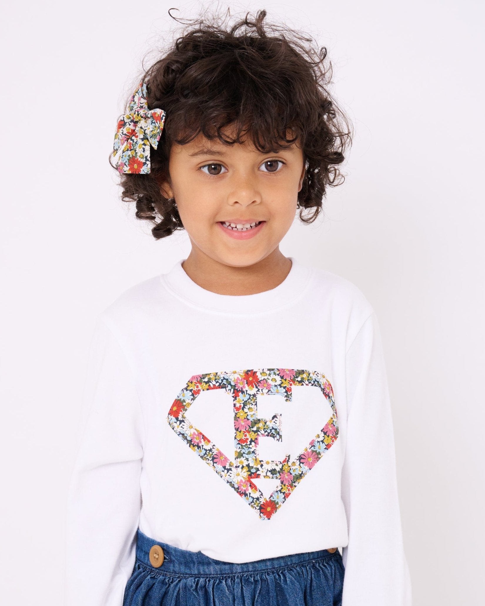 A 3 year old girl wearing a personalised white t-shirt with a superhero motif initial made from Libby floral Liberty print fabric.