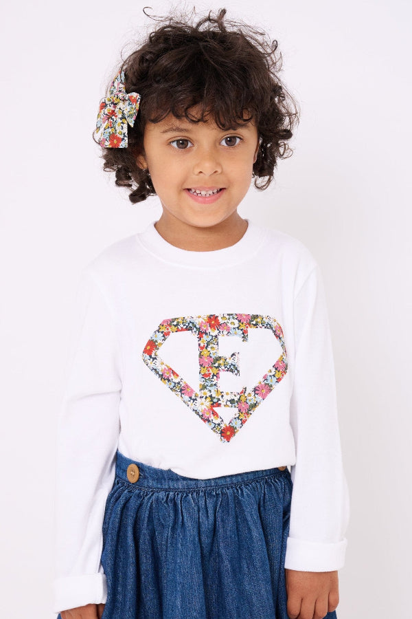 A 3 year old girl wearing a personalised white t-shirt with a superhero motif initial made from Libby floral Liberty print fabric.