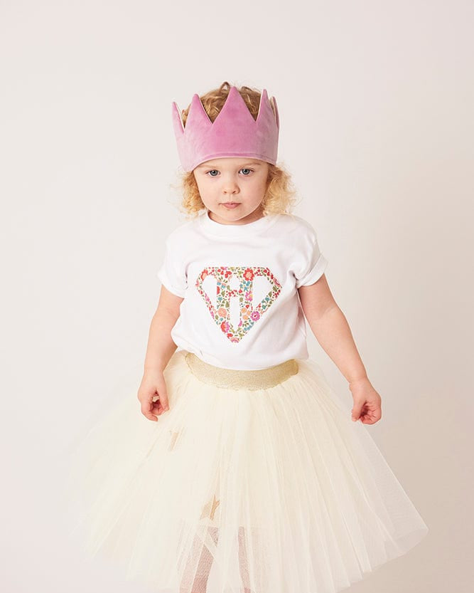 A 3 year old girl wearing a personalised white t-shirt with a superhero motif initial made from floral Liberty print fabric.
