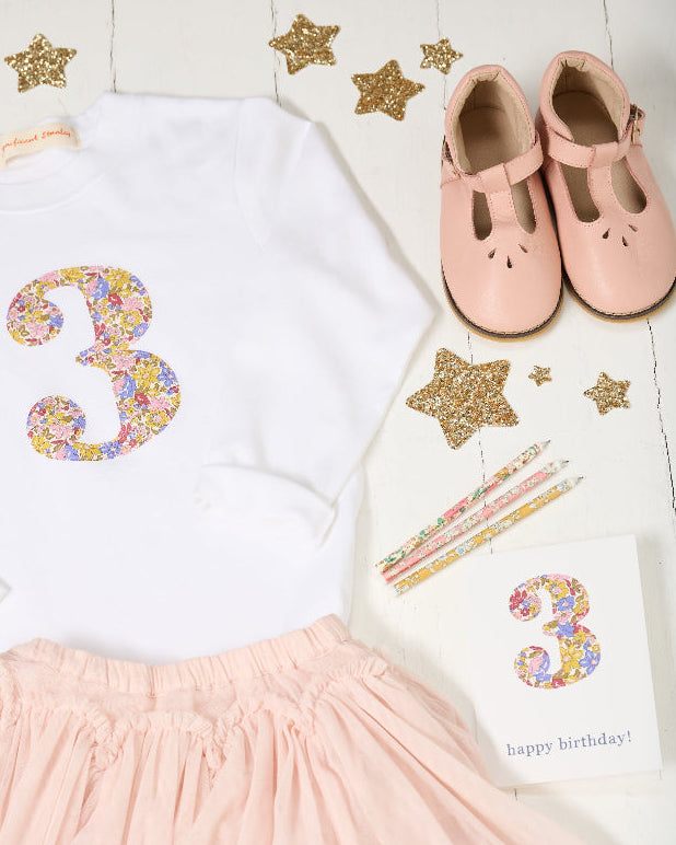 A children's birthday outfit set laid flat on a white wood background, including a white long-sleeve top with a large glittery pastel number 3, a light pink skirt, a pair of pale pink Mary Jane shoes, star confetti, pencils, and a small birthday card.