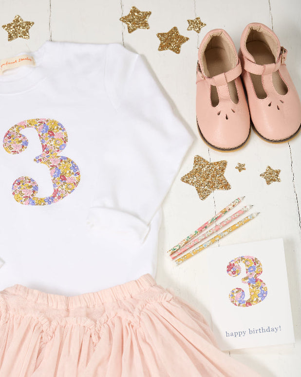 A children's birthday outfit set laid flat on a white wood background, including a white long-sleeve top with a large glittery pastel number 3, a light pink skirt, a pair of pale pink Mary Jane shoes, star confetti, pencils, and a small birthday card.