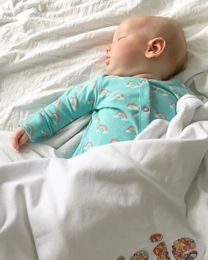A sleeping baby wearing a light blue raincloud-patterned bodysuit, lying on white bedding under a white blanket with the name 'Darcie' embroidered in Liberty Michelle floral pattern.