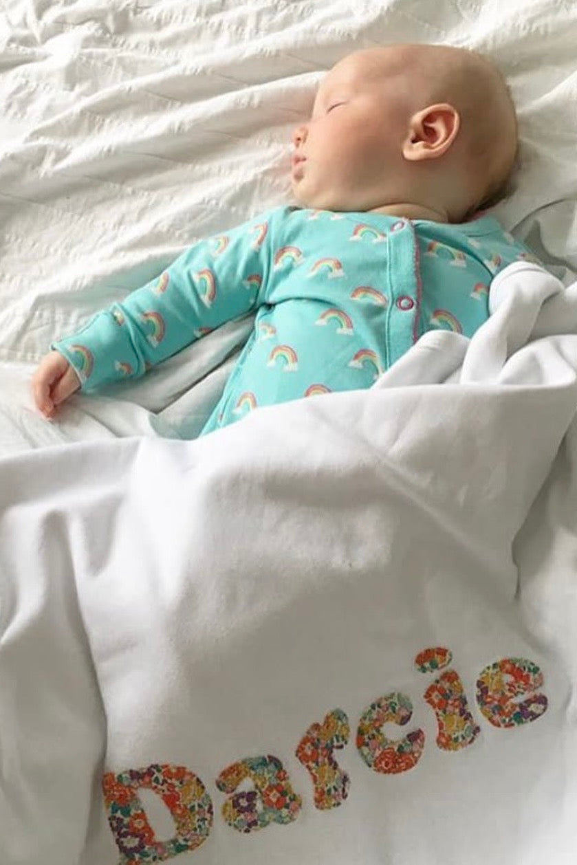 A sleeping baby wearing a light blue raincloud-patterned bodysuit, lying on white bedding under a white blanket with the name 'Darcie' embroidered in Liberty Michelle floral pattern.