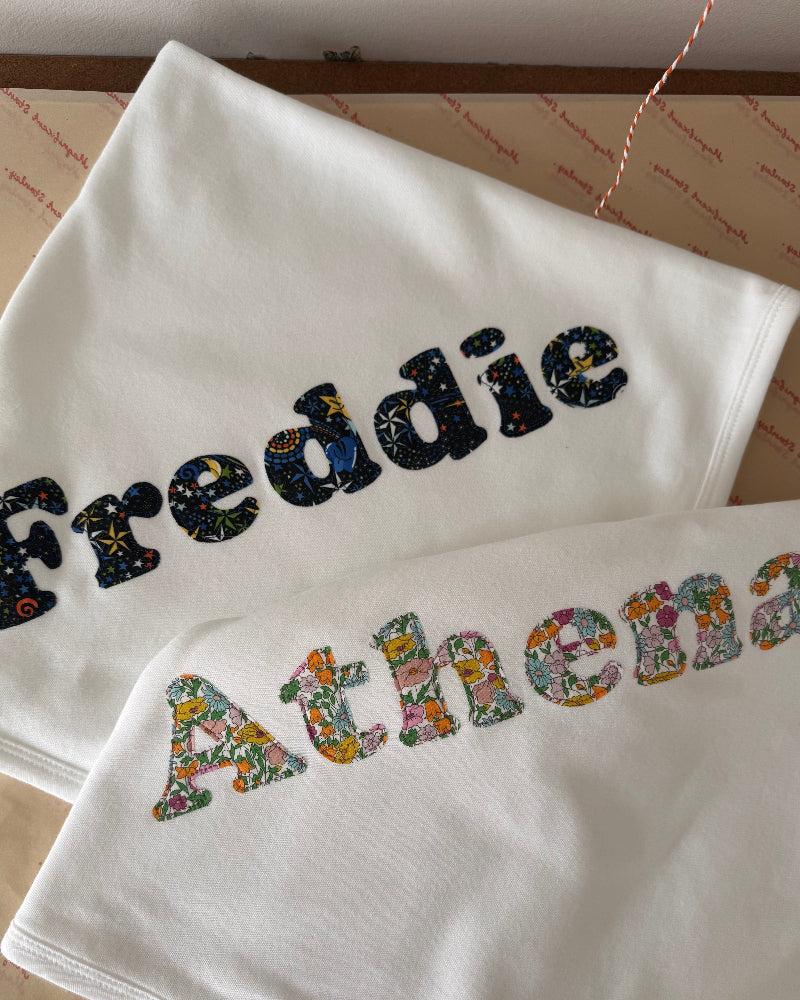 Two white baby blankets or swaddle blankets laid flat on a patterned surface. The top blanket is embroidered with the name 'Freddie' in dark Liberty space patterned letters, and the bottom blanket is embroidered with the name 'Athena' in multicoloured floral Liberty letters.