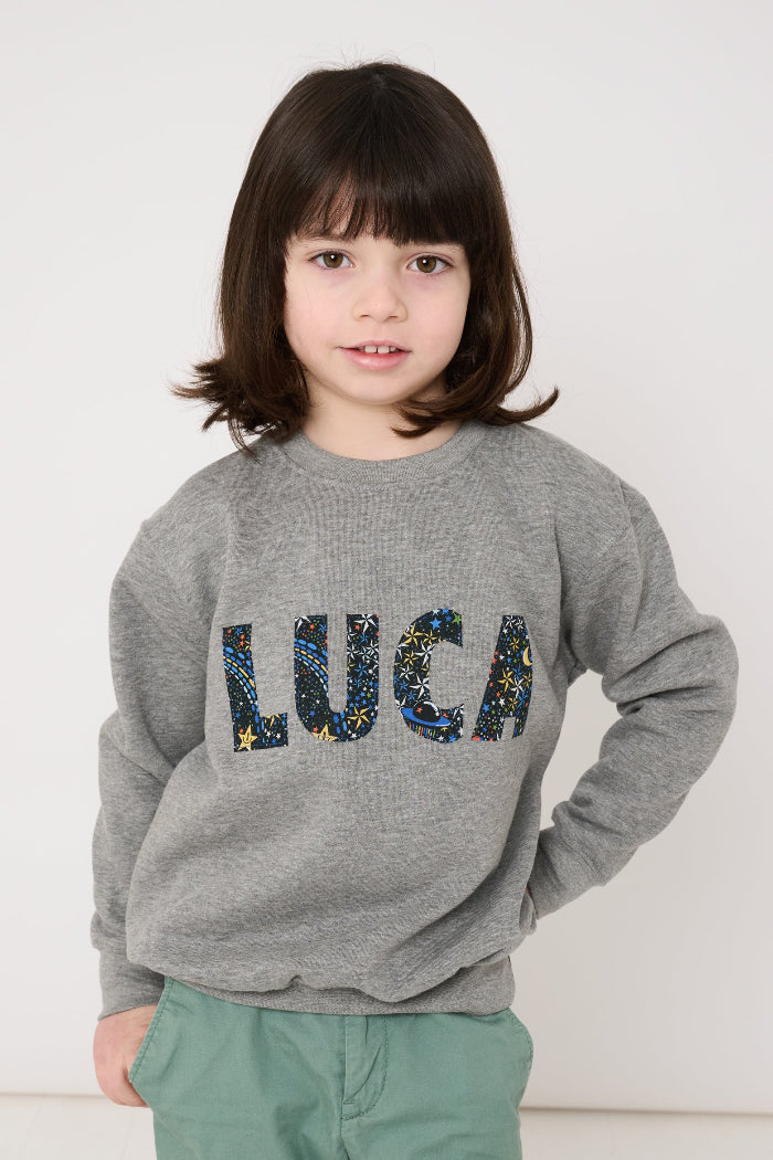 Child wearing a grey sweatshirt with 'LUCA' in colourful letters in space liberty print.