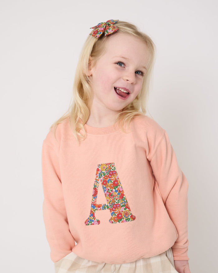 Young girl wearing a pink sweatshirt with a Liberty floral letter 'A' design and plaid trousers on a white background