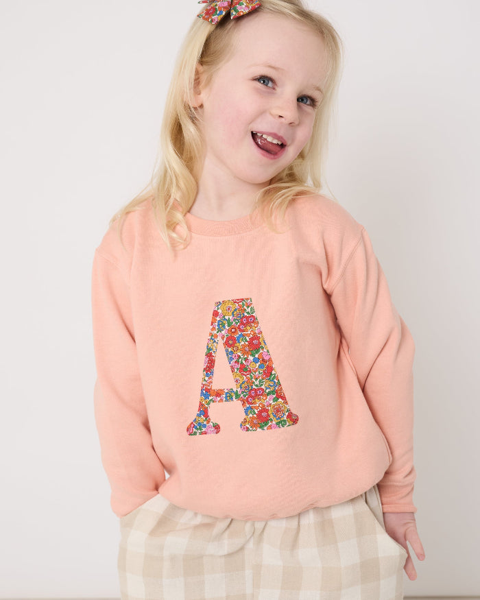 Young girl wearing a pink sweatshirt with a Liberty floral letter 'A' design and plaid trousers on a white background