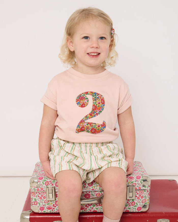 Child sitting on a red suitcase wearing a pink shirt with the number 2 and floral Liberty design, white shorts, and white shoes.