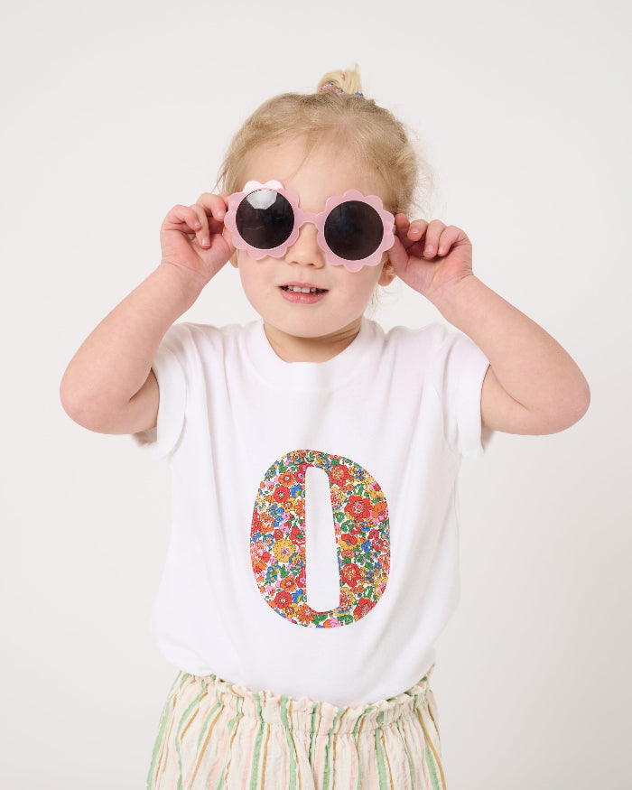 A 2 year old girl wearing a summery personalised white cotton t-shirt with her initial cut from Nell Annie and May floral liberty fabric.