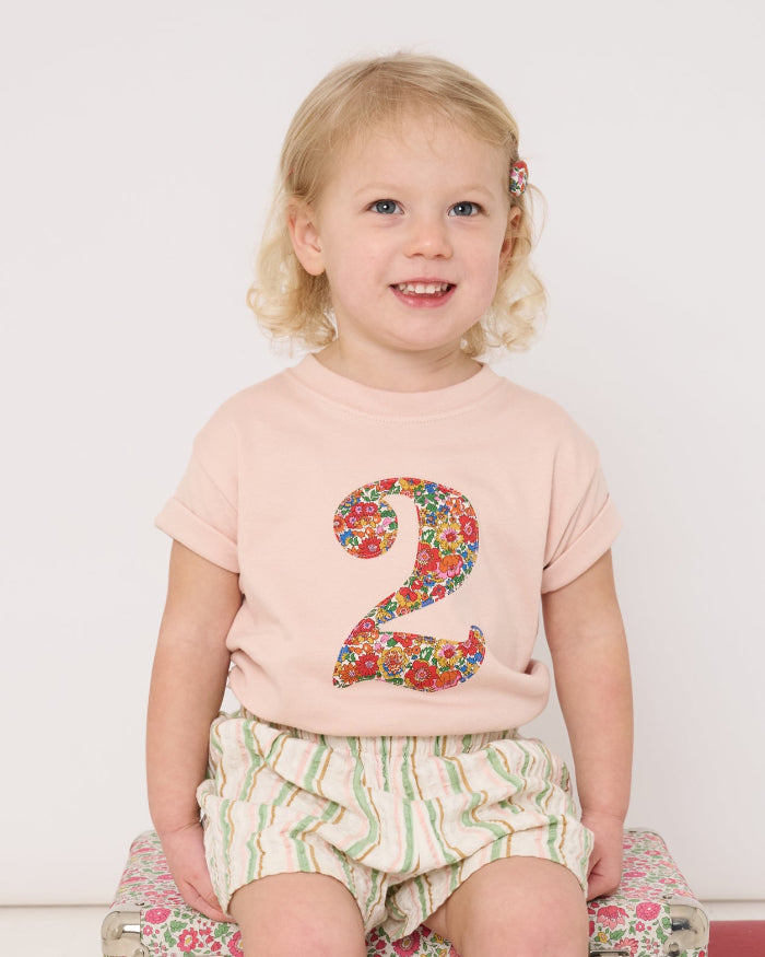 A 2 year old girl wearing a pale pink t-shirt with a Liberty Nell Annie and May floral number '2' on the front