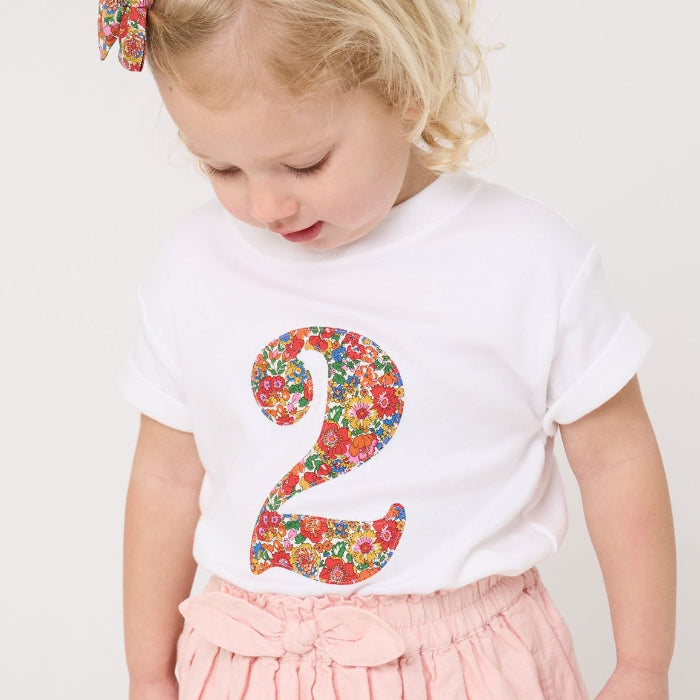 A 2 year old girl wearing a white cotton birthday t-shirt with the number 2 sewn on the front cut from liberty Nell Annie and May fabric.