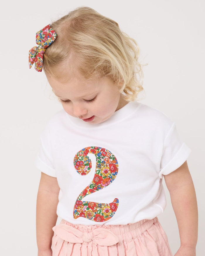 A 2 year old girl wearing a white cotton birthday t-shirt with the number 2 sewn on the front cut from liberty Nell Annie and May fabric.