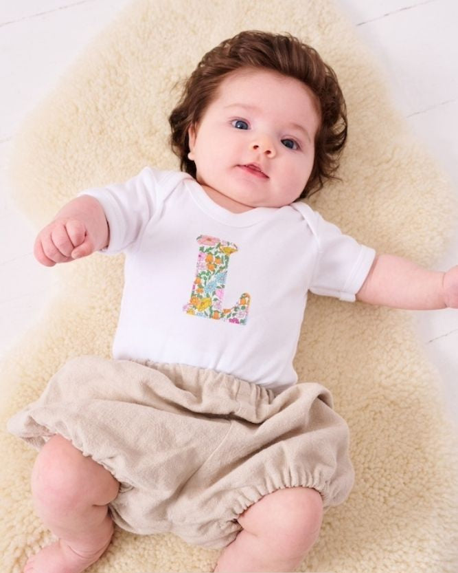 A smiling newborn baby wearing a personalised white bodysuit with an initial on the front cut from Liberty floral Poppy Forest fabric.