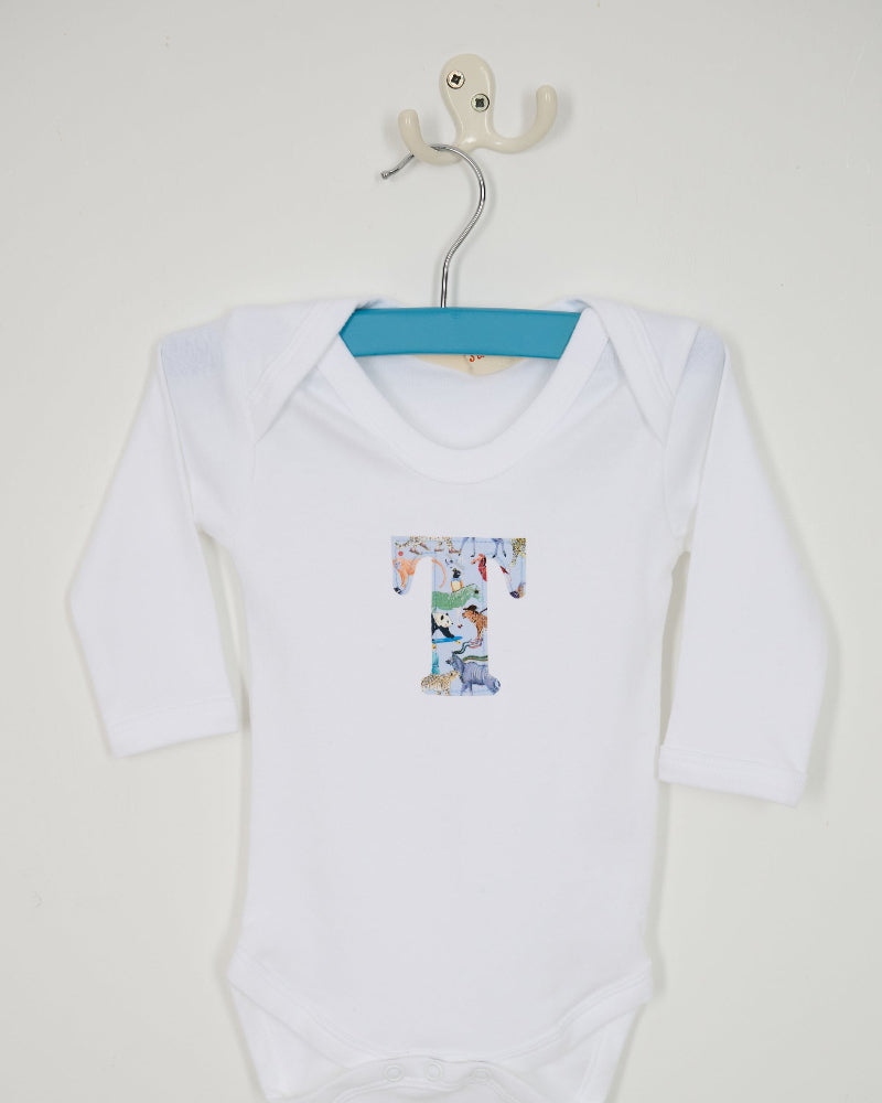 A white long-sleeve baby bodysuit hanging on a metal hanger, featuring a Quey 2 Liberty print graphic letter 'T' on the chest, set against a plain off-white wall.