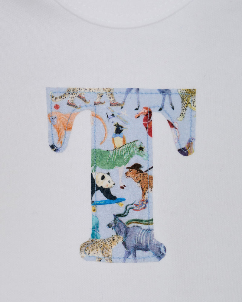 A close-up of a white baby grow personalised with a large letter 'T' printed with a Liberty quey 2 safari animal pattern, including a zebra, giraffe, panda, and cheetah, on a white background.