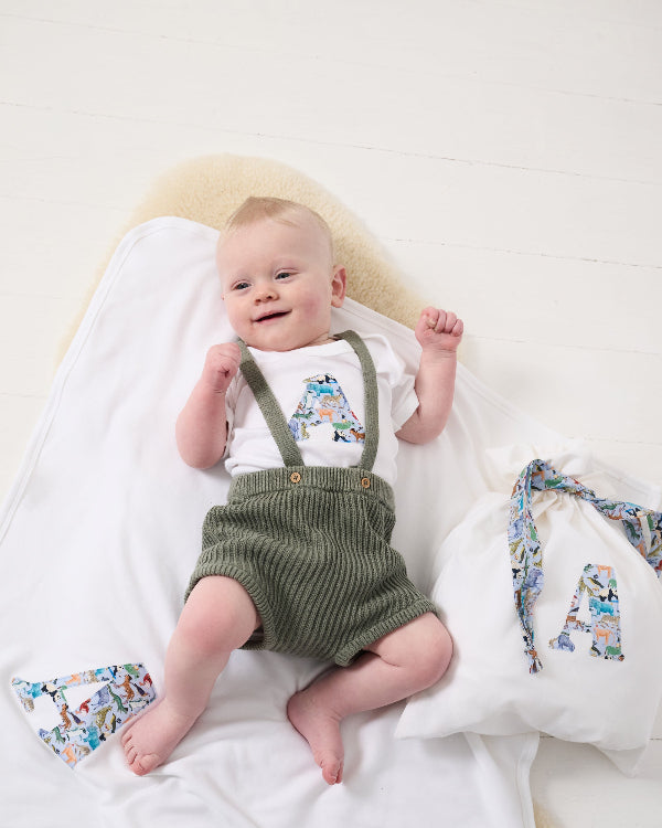 A newborn baby laying on a Personalised Cotton Baby Blanket with their initial sewn on it made from zoo animal liberty print. worn with matching babygro and gift bag.