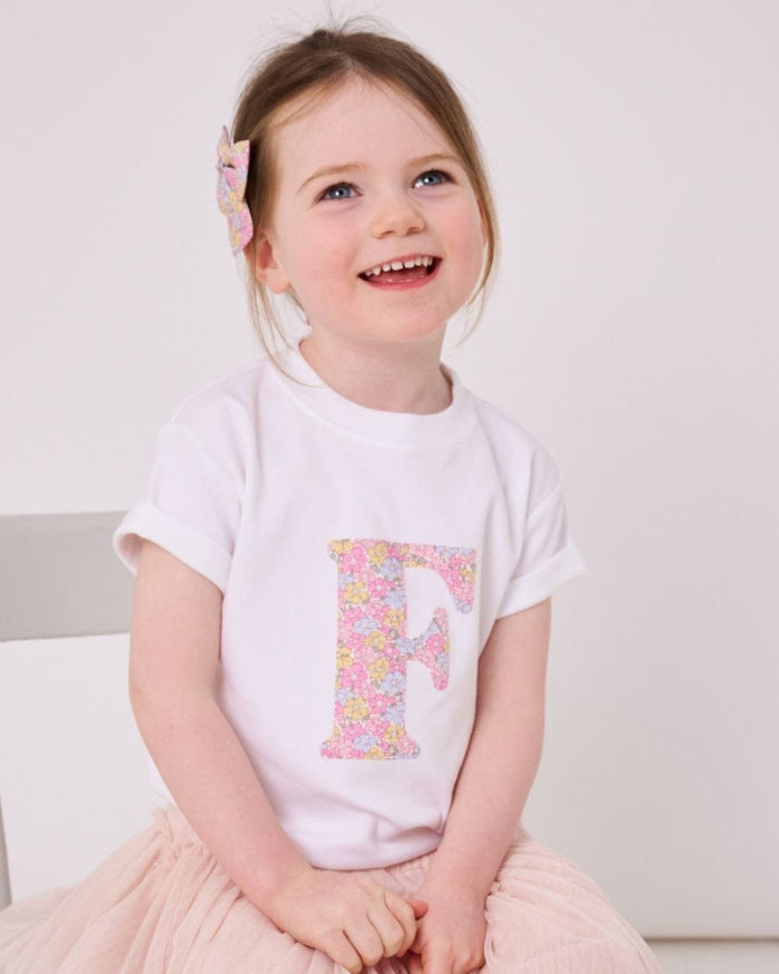 A 5 year old girl wearing a white personalised cotton t-shirt with her initial on the front in Sadie Rose Liberty fabric.
