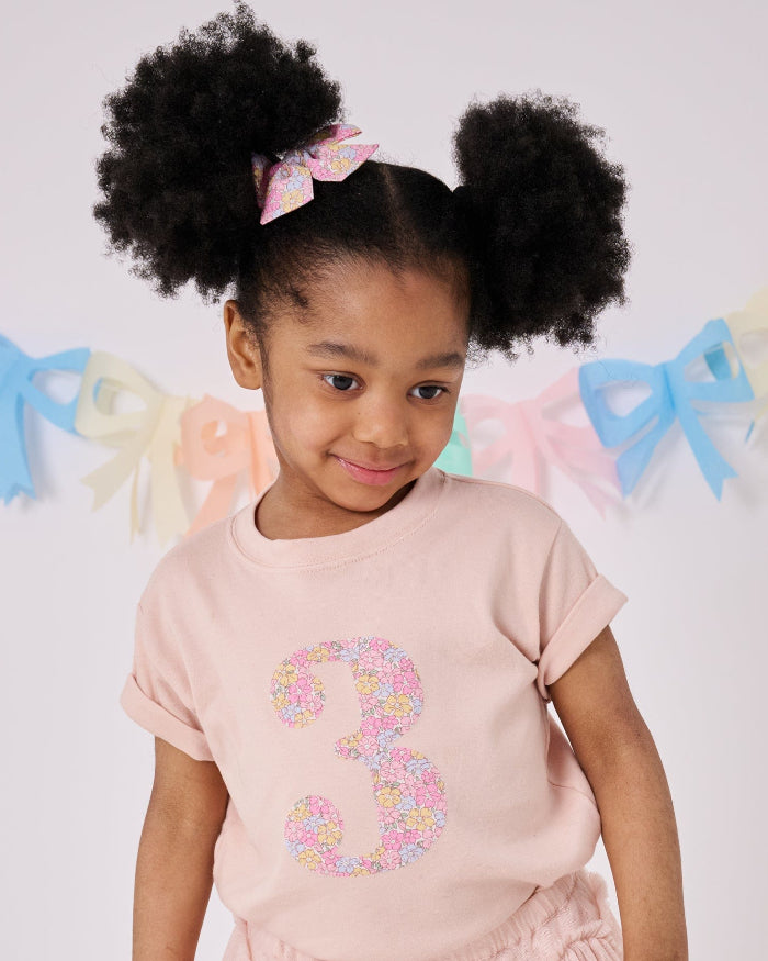 A child wearing a blush pink short sleeve t-shirt with the number 3 hand-appliqued in a floral Liberty Print.