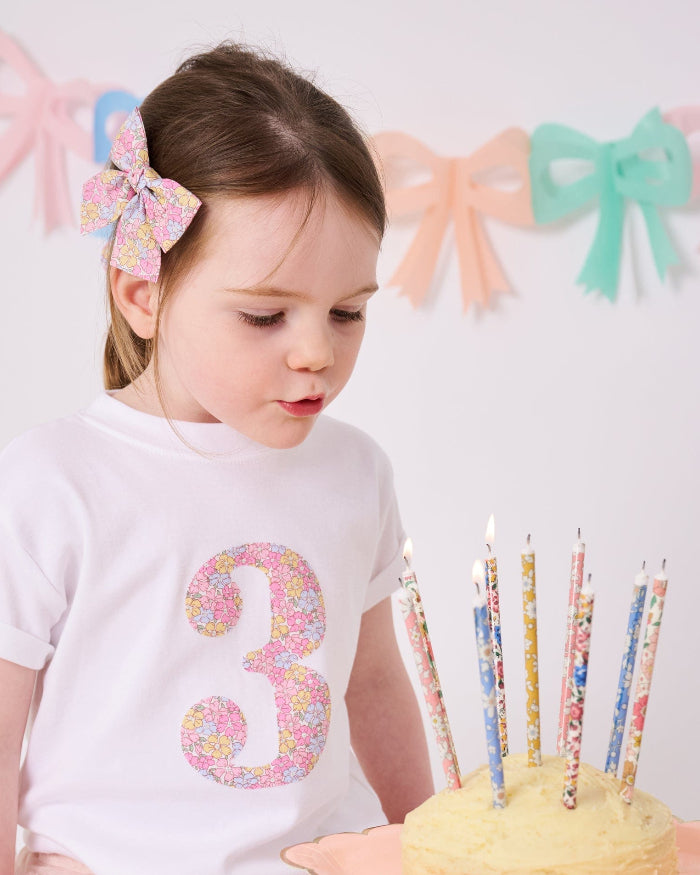 a 3 year old girl on her birthday wearing a white t-shirt with a number 3 cut from Sadie Rose Liberty floral fabric on the front. She is blowing out candles on a birthday cake with liberty candles