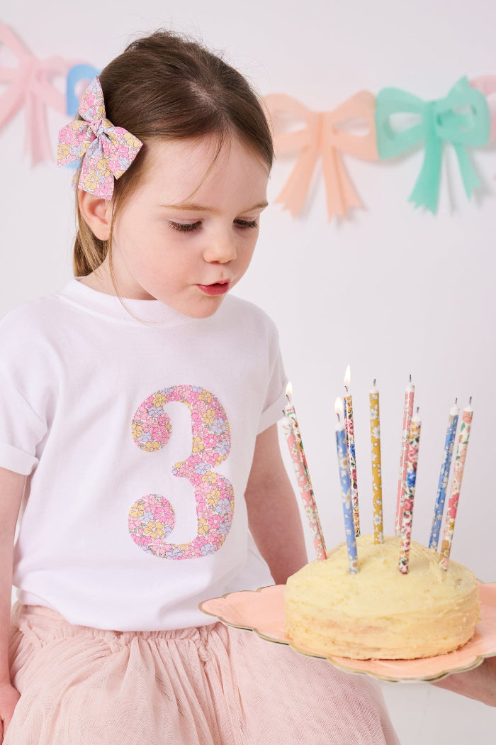 a 3 year old girl on her birthday wearing a white t-shirt with a number 3 cut from Sadie Rose Liberty floral fabric on the front. She is blowing out candles on a birthday cake with liberty candles