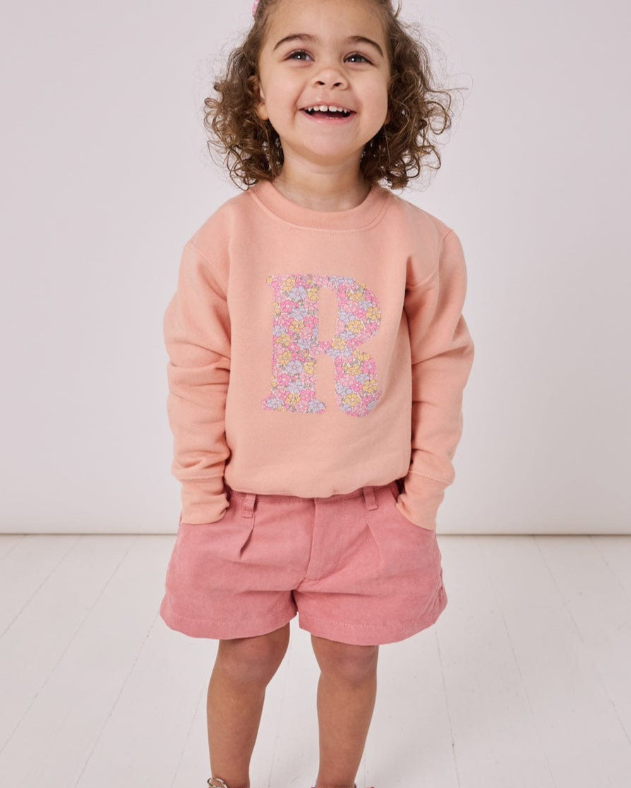 A smiling 2 year old girl wearing a personalised dusty pink sweatshirt with her initial on in floral Sadie Rose Liberty print on the front.
