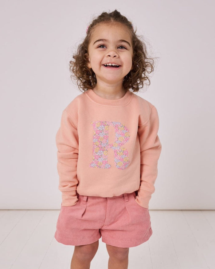 A smiling 2 year old girl wearing a personalised dusty pink sweatshirt with her initial on in floral Sadie Rose Liberty print on the front.