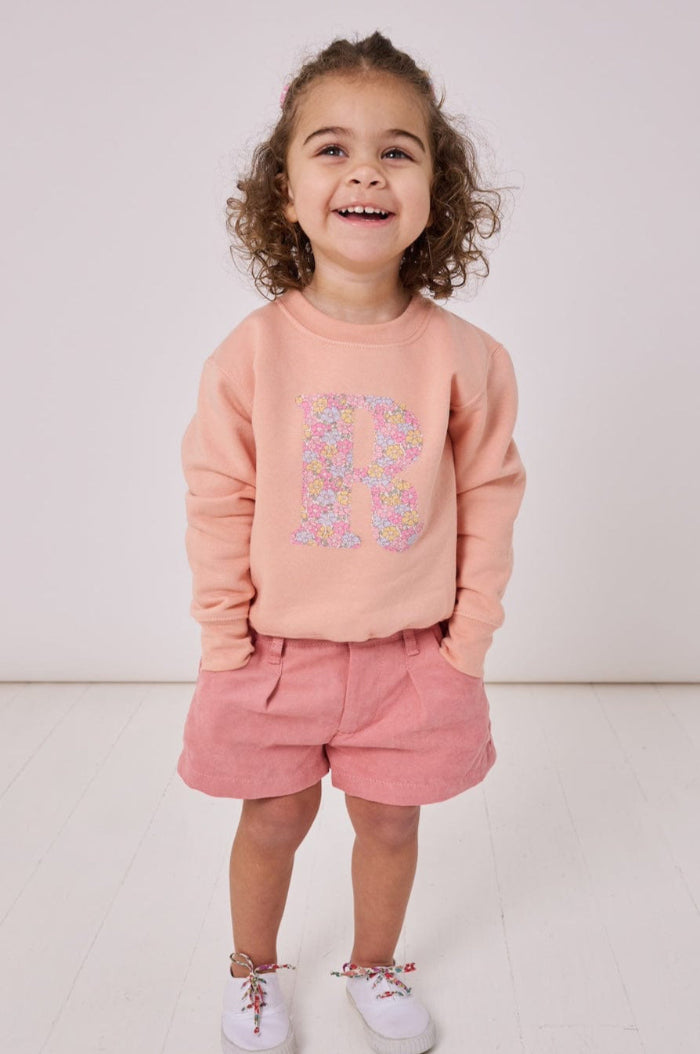 A smiling 2 year old girl wearing a personalised dusty pink sweatshirt with her initial on in floral Sadie Rose Liberty print on the front.