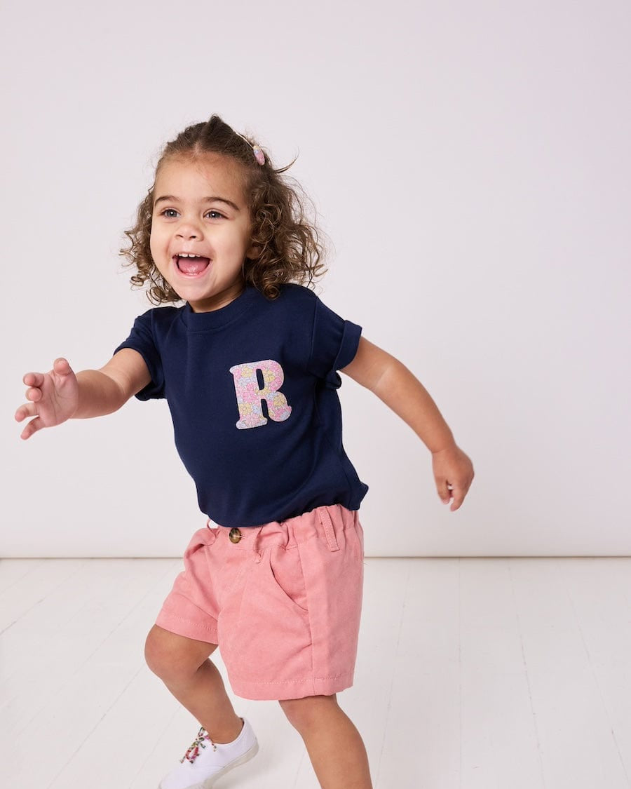 A smiling 2 year old girl wearing a personalised kids cotton t-shirt with a Liberty print initial on the front.