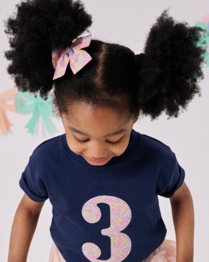 A 3 year old birthday girl wearing a navy cotton kids birthday t-shirt with the number 3 in Sadie Rose pink floral Liberty print on the front.