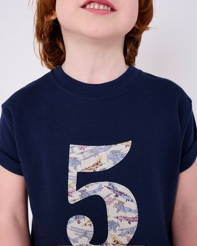 a 5 year old boy wearing a navy birthday t-shirt with a number 5 cut from Tom's Jet plane liberty print on the front