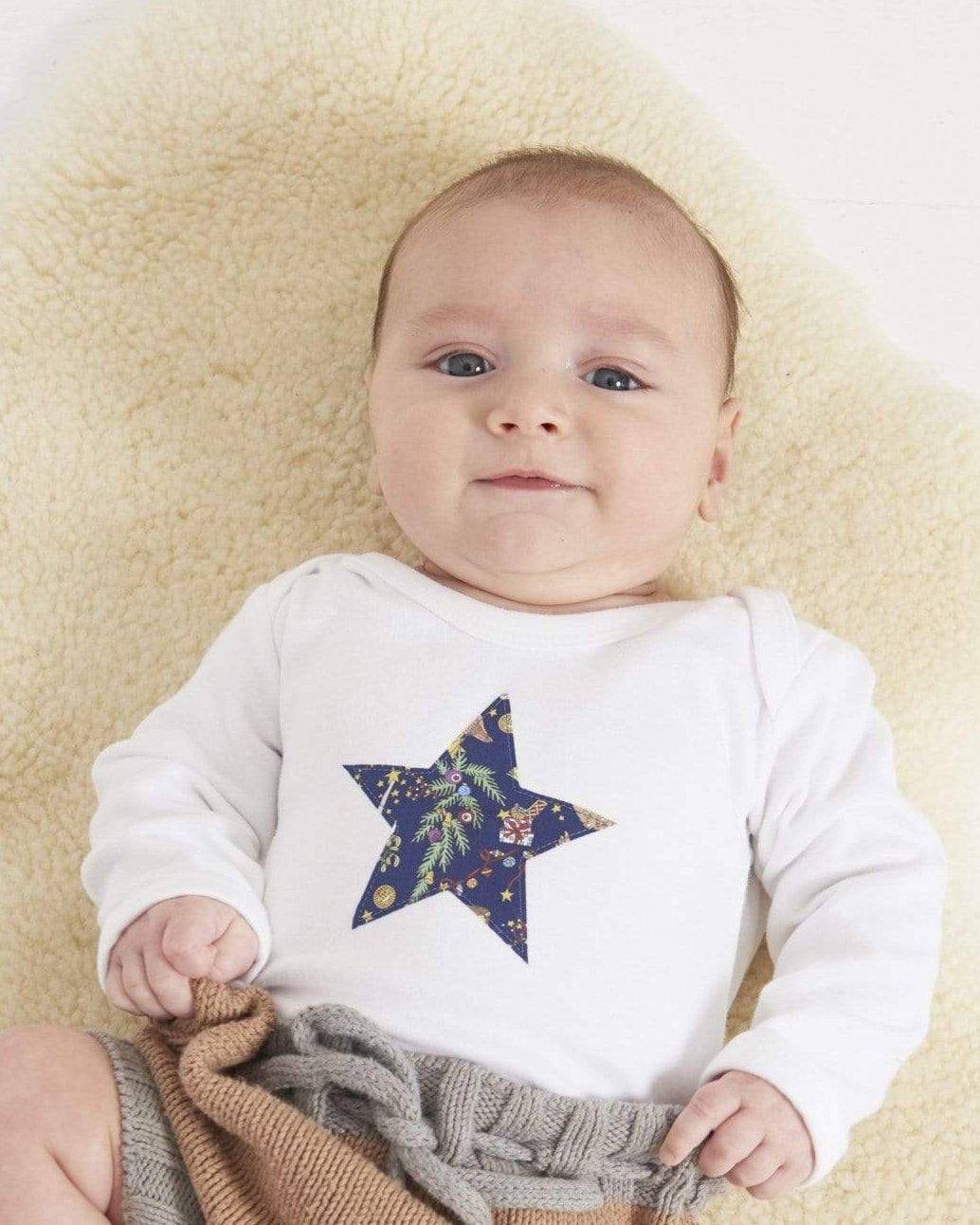 A baby wearing a white bodysuit babygrow with a Liberty Christmas print festive star sewn on the front.