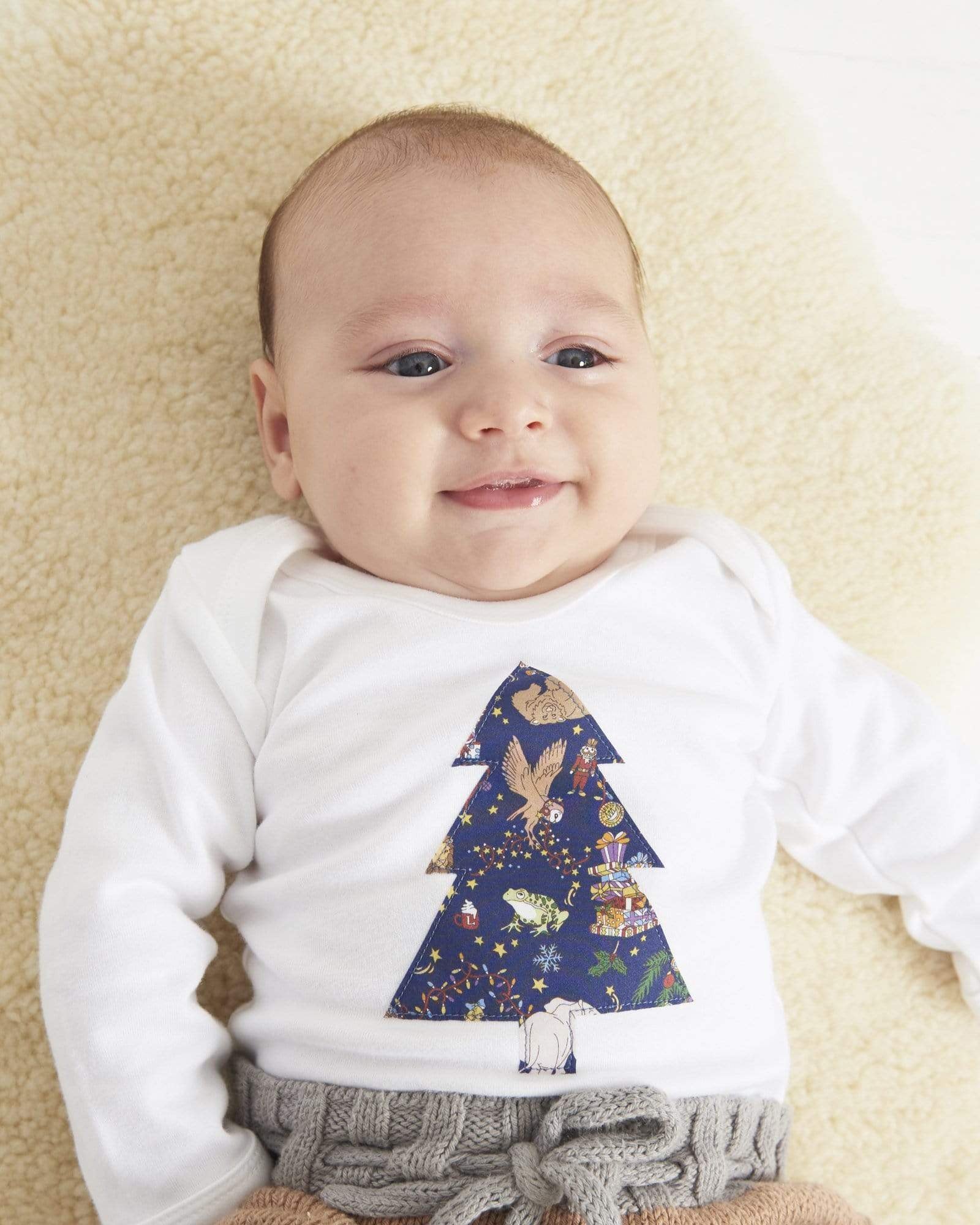 A baby wearing a white bodysuit babygrow with a Liberty Christmas print Christmas tree sewn on the front.
