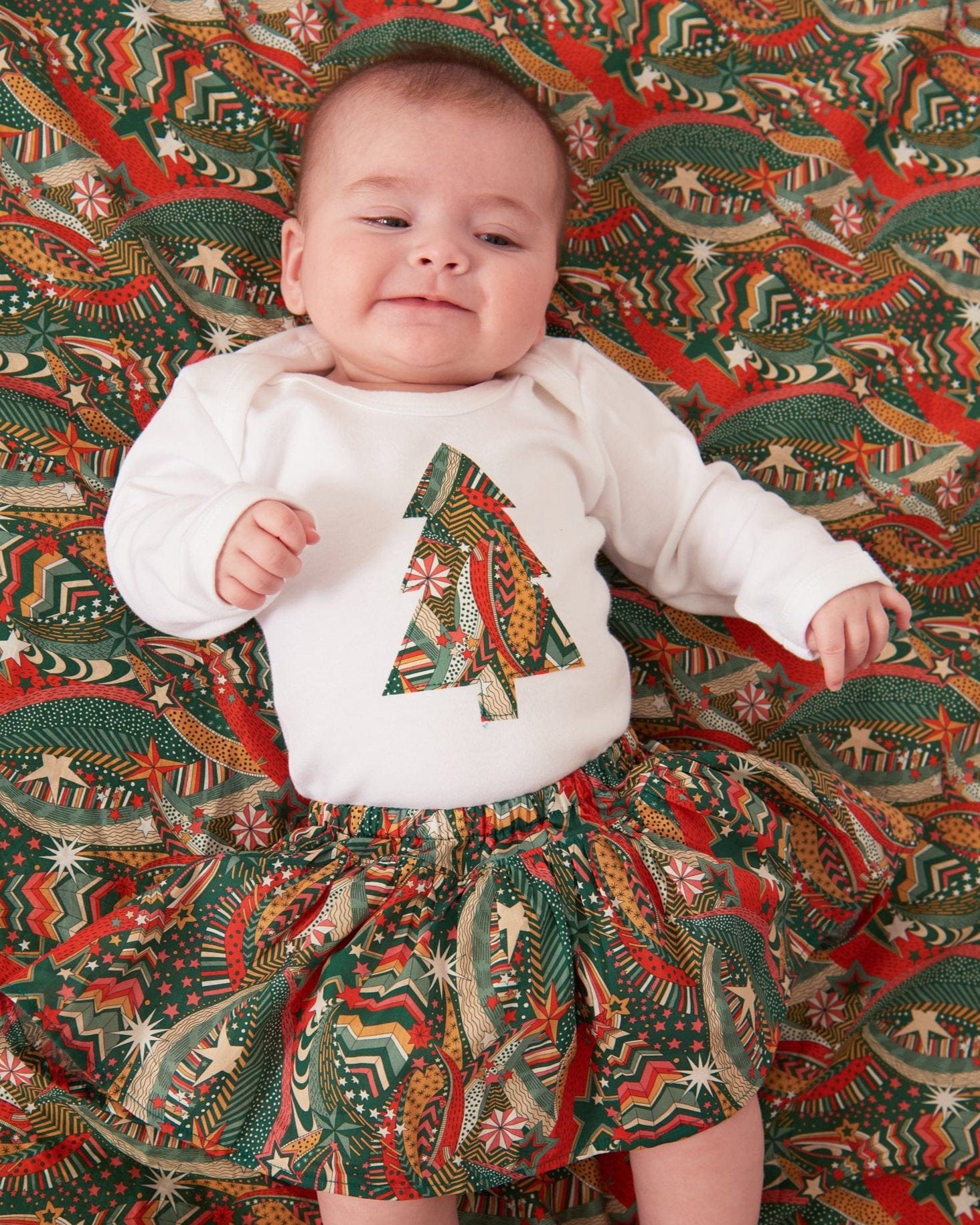 A baby wearing a white bodysuit babygrow with a Liberty My Little Star print festive Christmas tree sewn on the front.