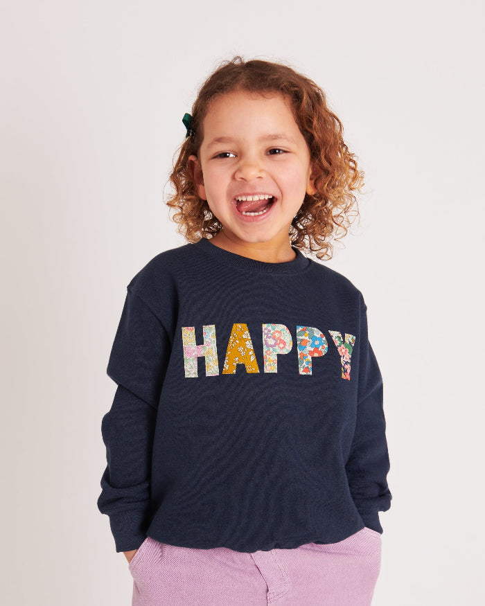 A 5 year old girl wearing a Magnificent Stanley navy sweatshirt with the word HAPPY Mixed Liberty Print sewn onto the front