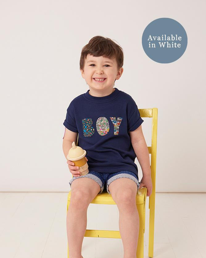 A little boy wearing a navy t-shirt with a name on in mixed Liberty fabrics.