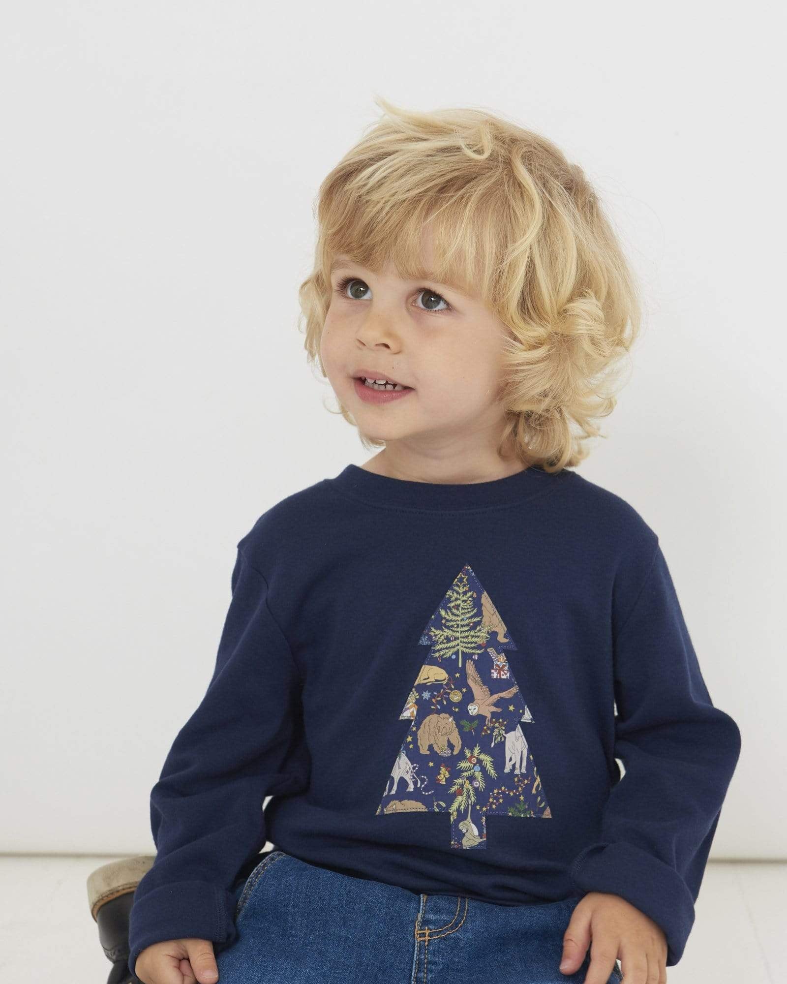A 3 year old boy wearing a navy long sleeve Christmas t-shirt with a Christmas Tree on the front made from Liberty Christmas print .