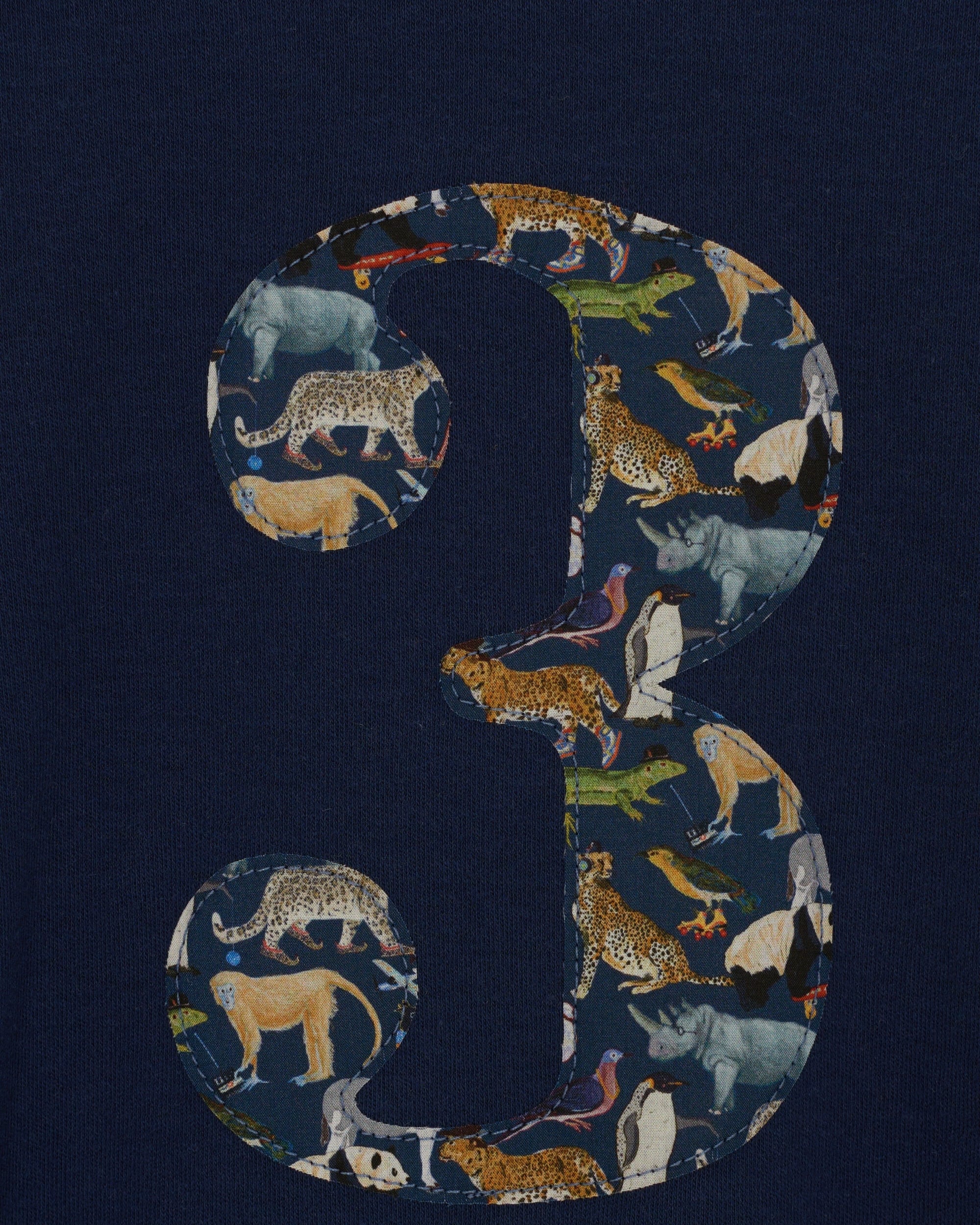 A 3 year old boy wearing a navy long sleeve t-shirt with a  number 3 on the front on in Liberty Quey Zoo animal print fabric. A close up of the appliqué stitching.
